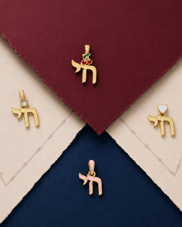 Four gold-tone Hebrew letter pendant charms arranged on a styled flat-lay background of deep burgundy, cream, and navy textured paper. The charms include small colorful enamel accents in green, red, pink, and white.